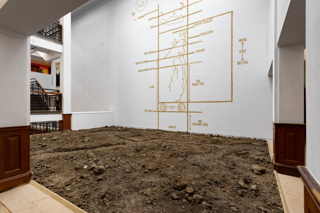 An atrium with white walls and large open archways. In the centre is a square bed of clay and earth. Painted on the wall is a diagram showing a cross section of the Earth marking space and time.