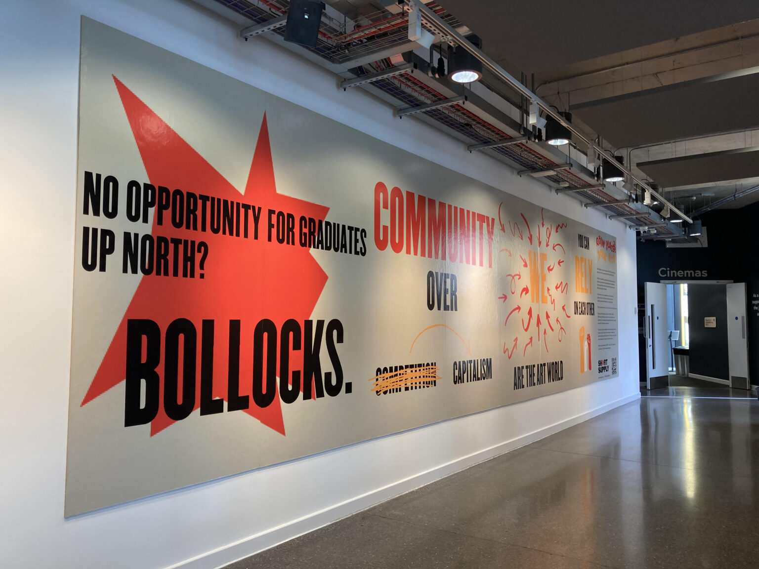 A billboard on a white wall contains a number of slogans, including 'No opportunity for graduates up North? Bollocks!'.