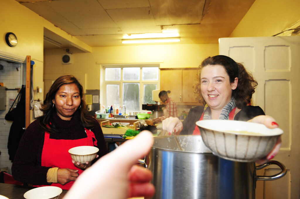 A photo of a person handing over a bowl of soup to the person behind the camera while another person looks on in a soup kitchen.