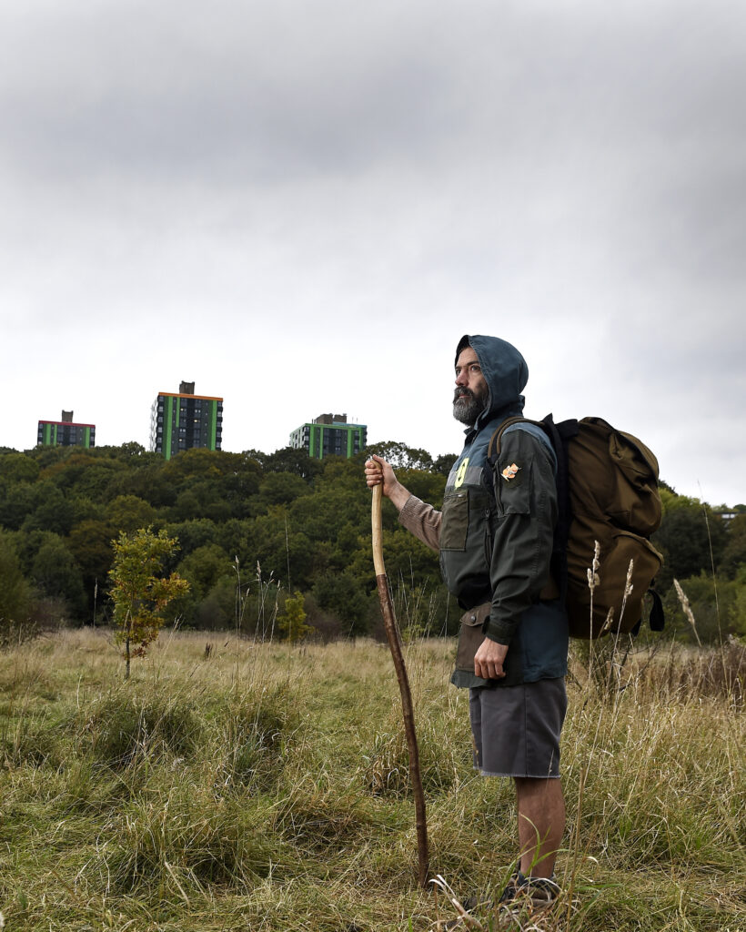 A photo of a man with a rucksack and walking stick stands in a a field with a grey sky in the background.