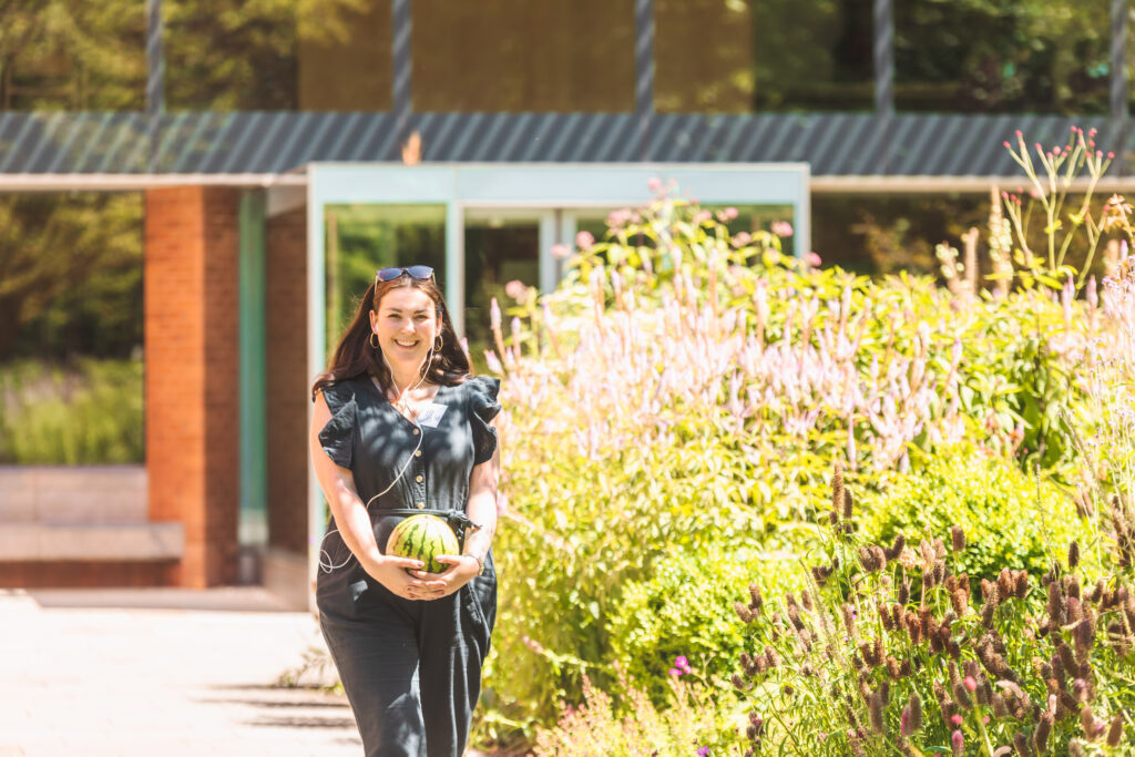 A smiling woman walks in a sunny park holding a watermelon at her lower abdomen.