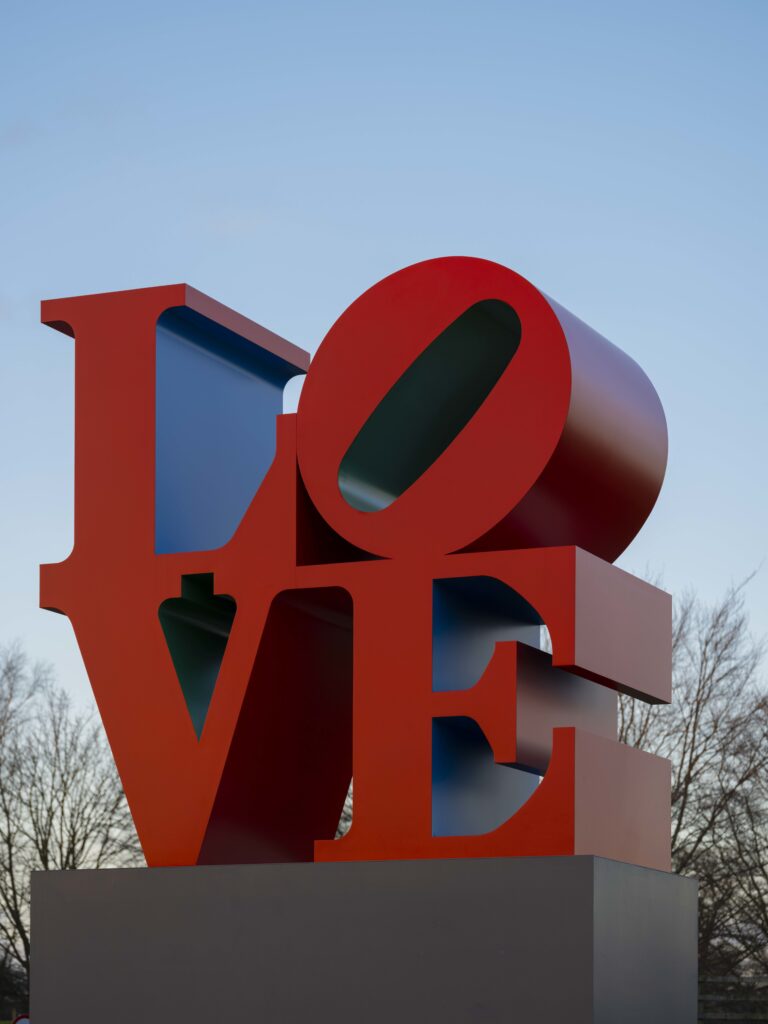 A red, blue and green sculpture spelling 'LOVE'. The letters 'L' and 'O' are stacked on top of 'V' and 'E'. The O is slanted, leaning to the right.
