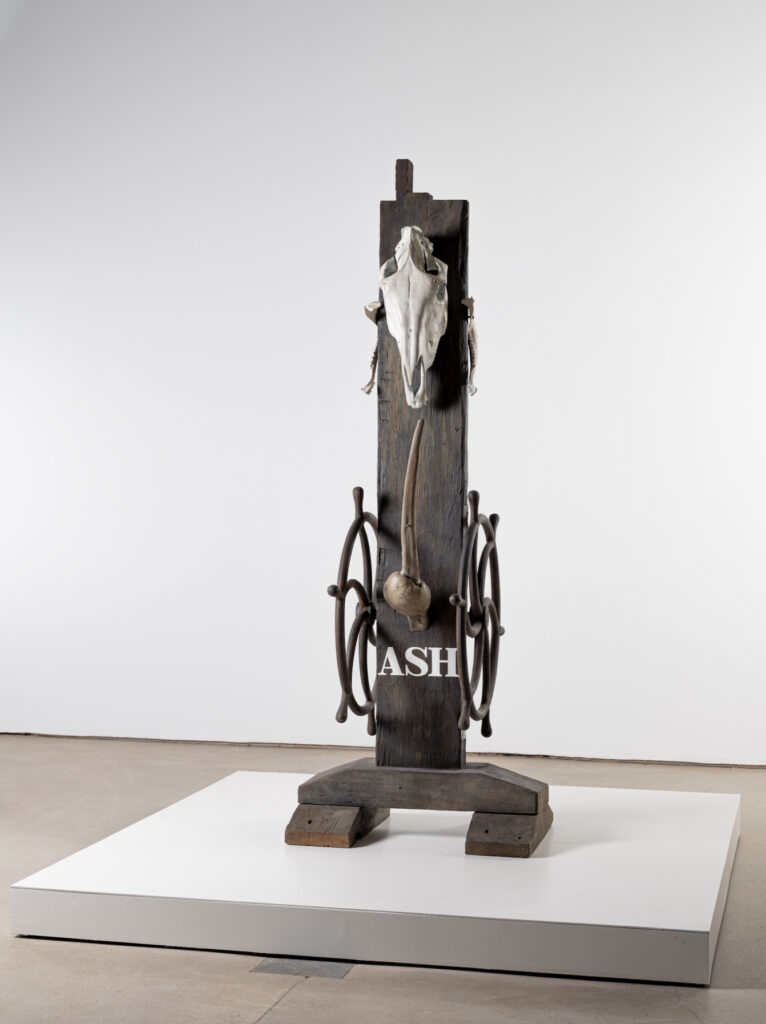 A tall, vertical sculpture made of wood, bones, an animal skull and metal wheels. The word 'ASH' is stencilled in white. The work is cast in bronze, but the textures appear life-like.