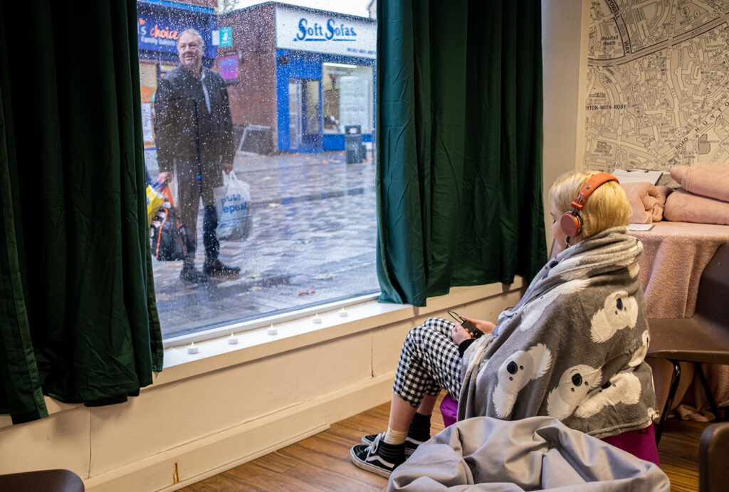 A young person sits on a beanbag listening to something on headphones in front of a window, an older person in the street outside looking through
