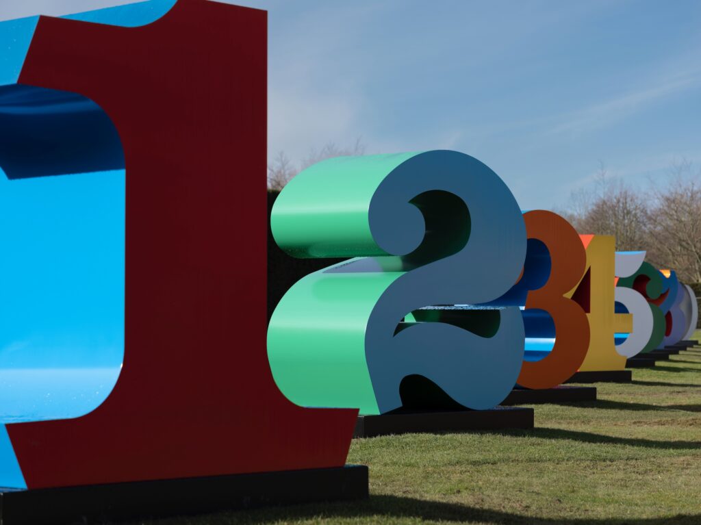 Large aluminium number sculptures, in bright colours, counting up from one. The sculptures are placed on grass with blue sky in the background.