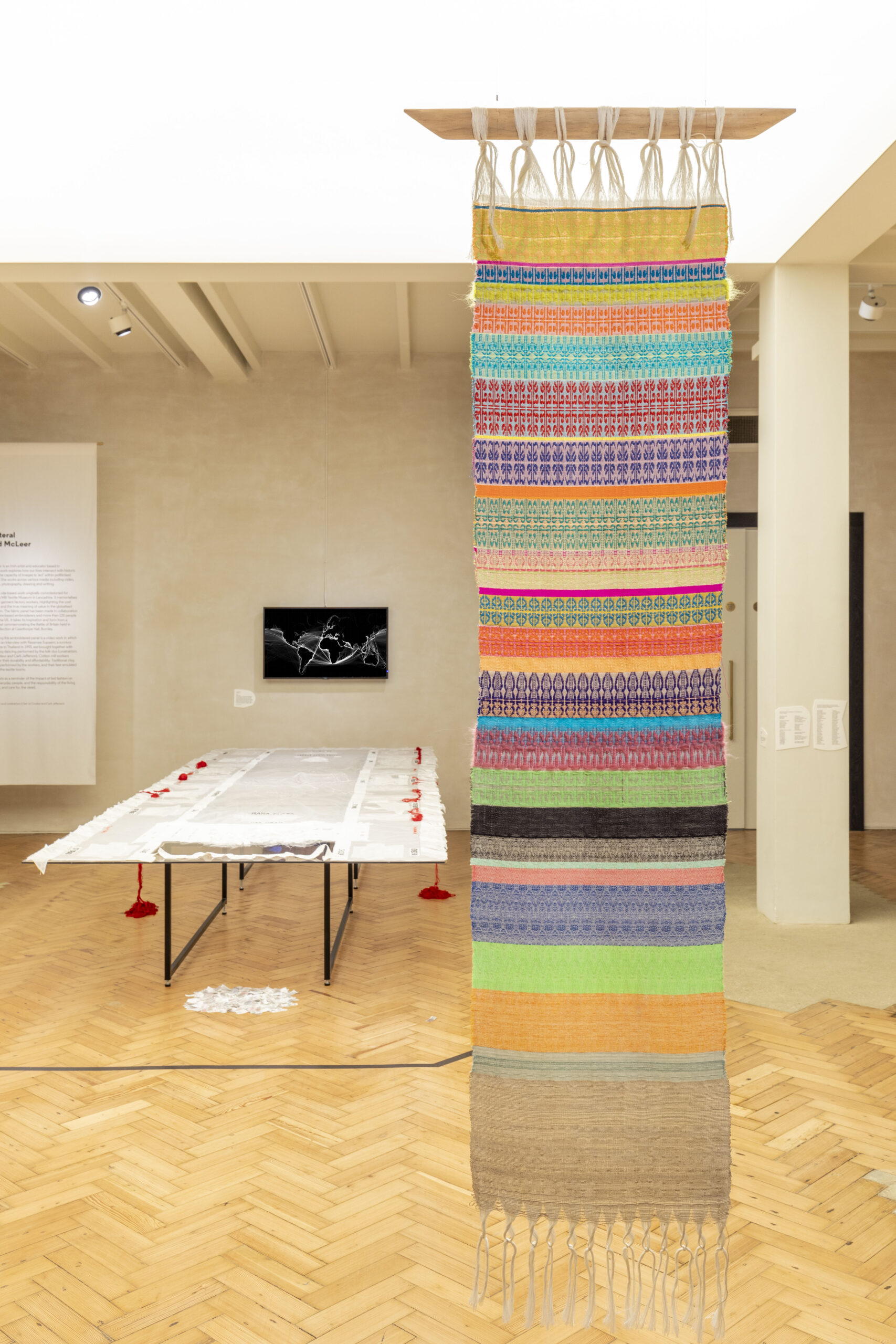 An installation view of an exhibition of textile-based works including a hanging colourful weaved garment and a table display of fabrics, in a brightly lit space with parquet flooring.