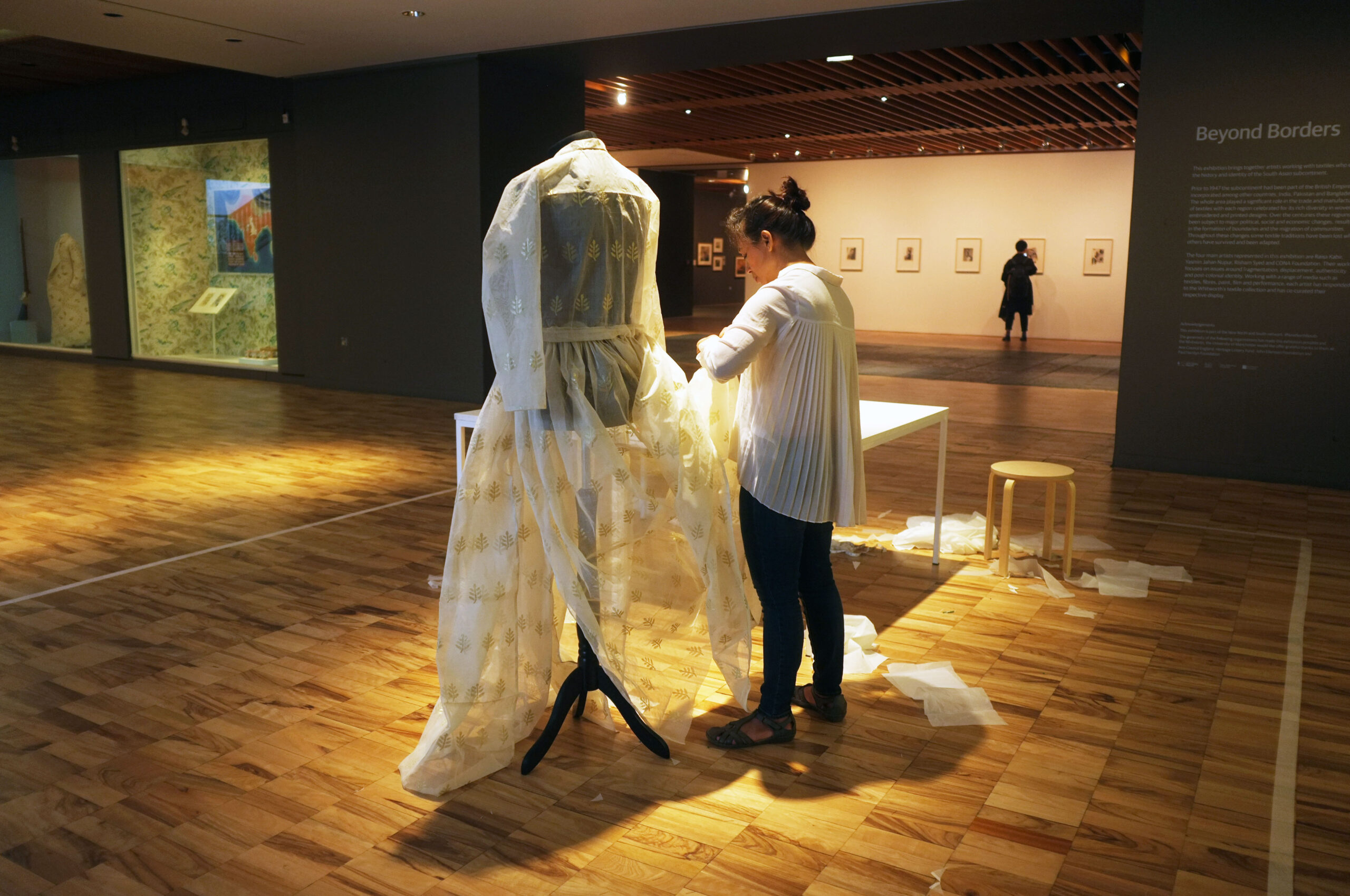 A woman stands sewing a white sheer dress on a mannequin stand in a darkened gallery space under a spotlight.