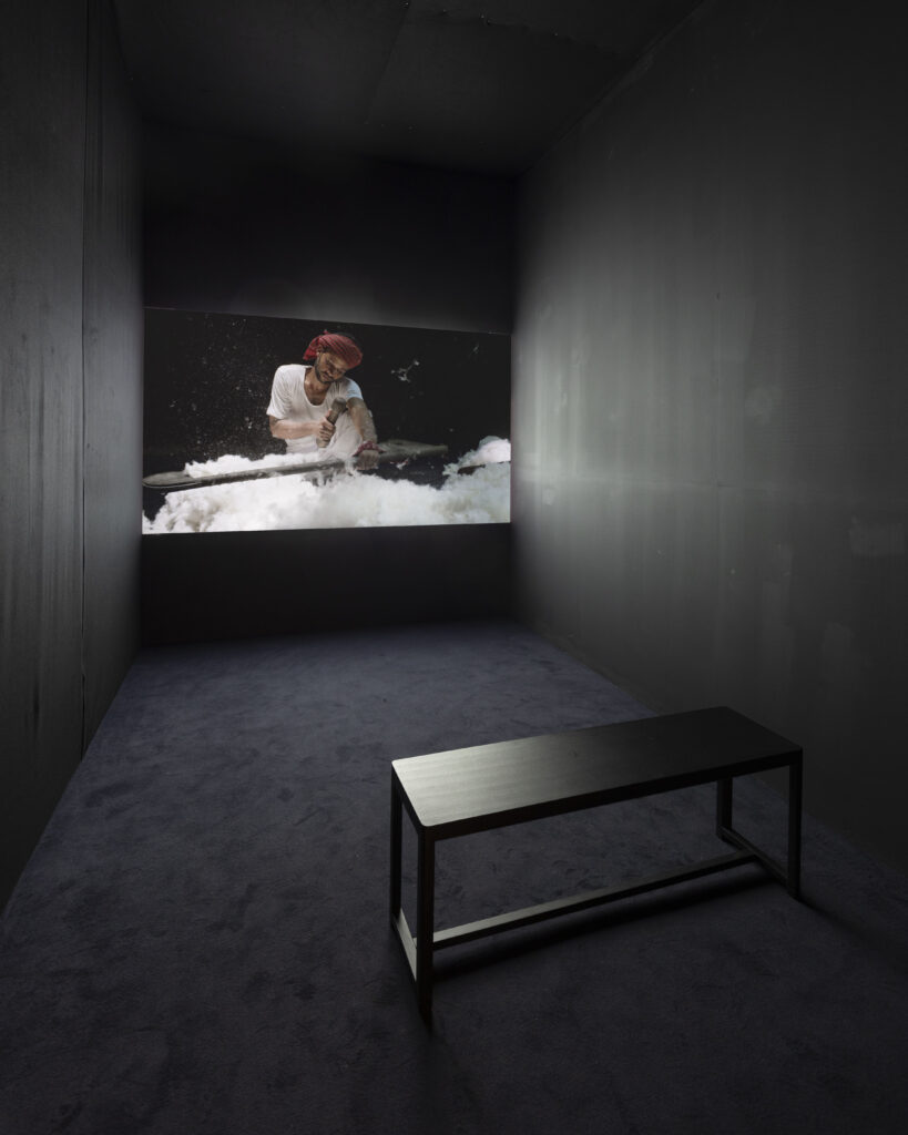 A video of a person processing cotton being screened in a dark room with a bench in the foreground.