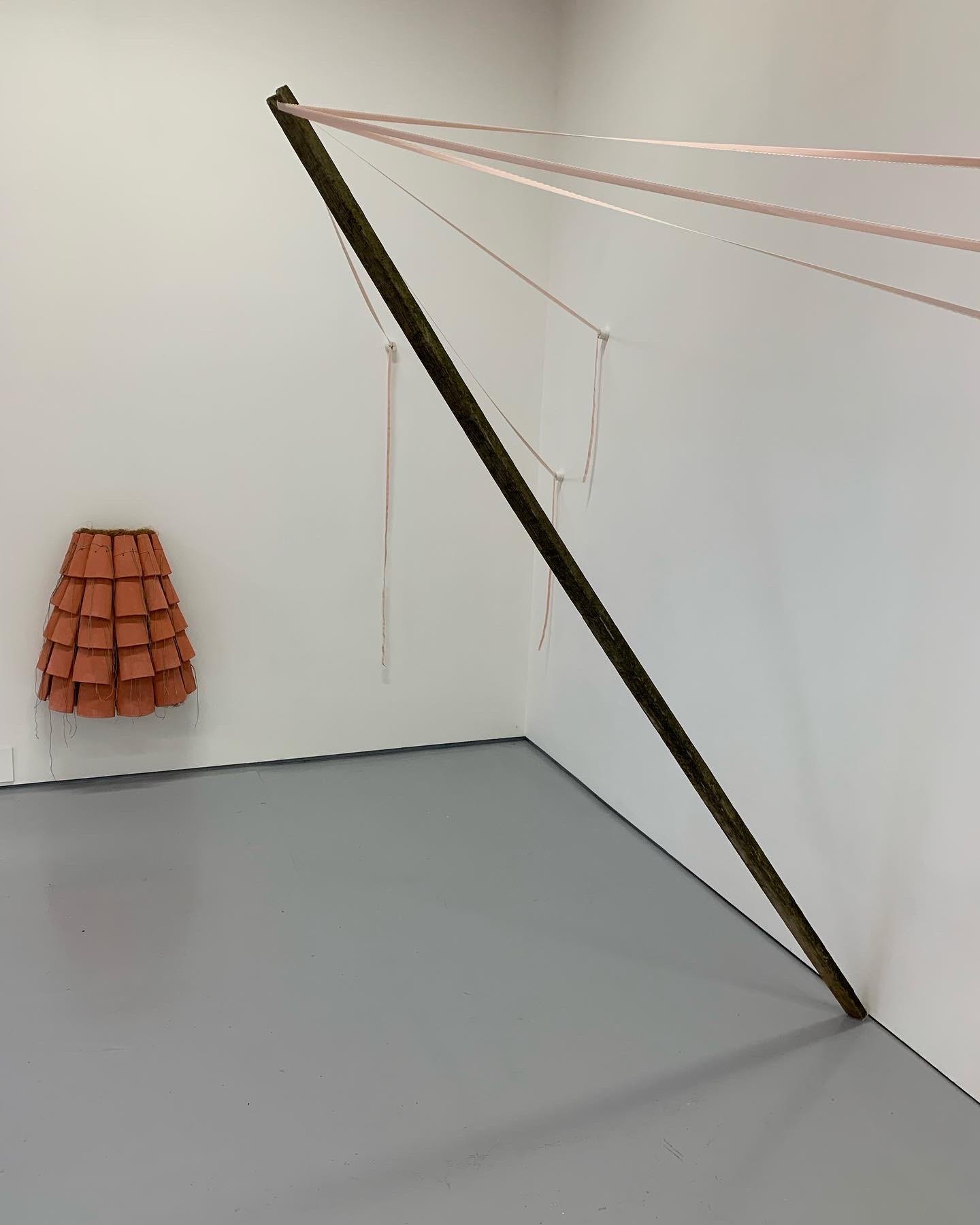 A wooden stick is held at an angle from the corner of a white wall, suspended from the top by lengths of peach coloured bra strap