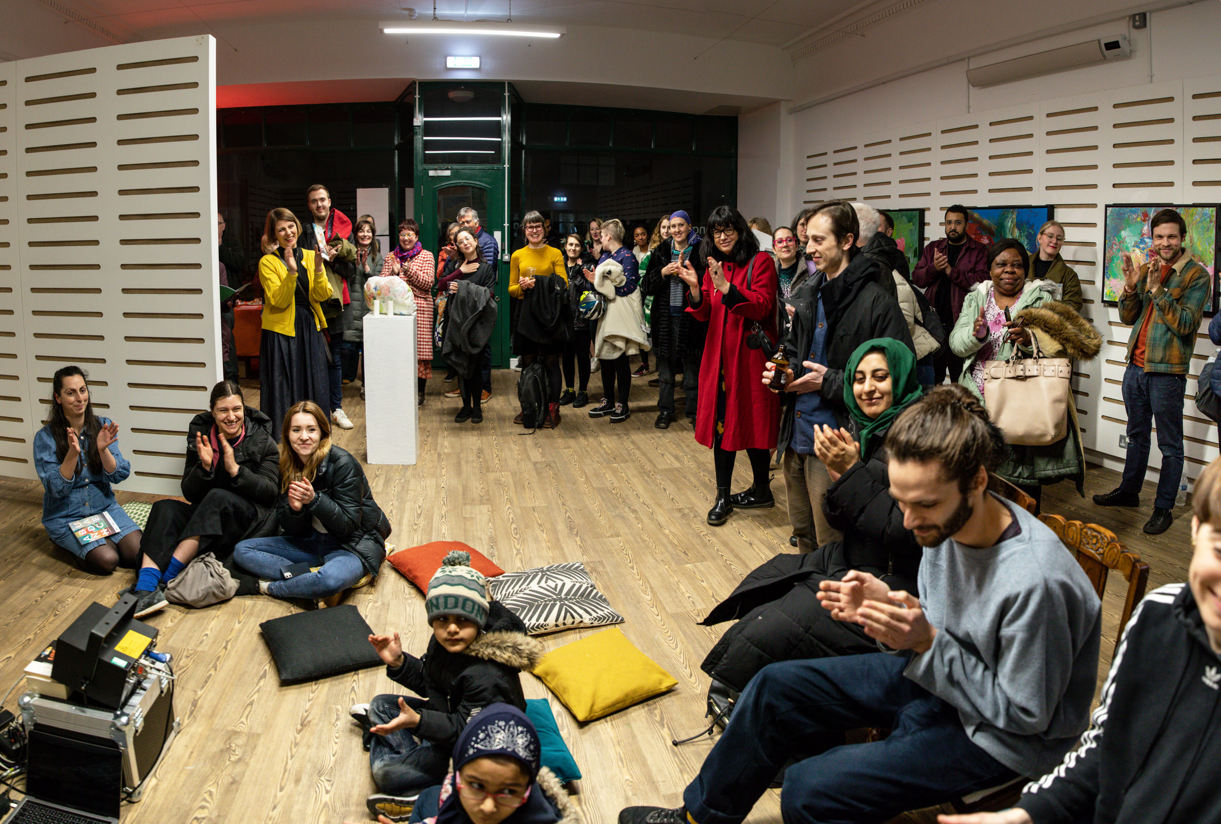 A gallery space full of people standing around the edges, some sitting on cushions on the floor. There are people of all ages and genders and colours. Everyone is applauding and smiling.