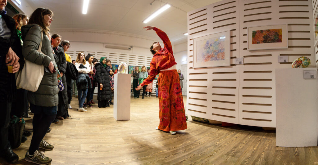 A dancer in orange clothing with one arm raised overhead, surrounded by people watching