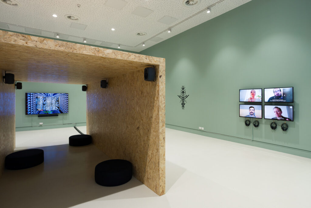 A photograph showing a view of the gallery. The walls are painted green and several screens showing videos are visible. A plywood tunnel in the centre of the room has cushions so visitors can sit within it, and speakers playing an audio work.