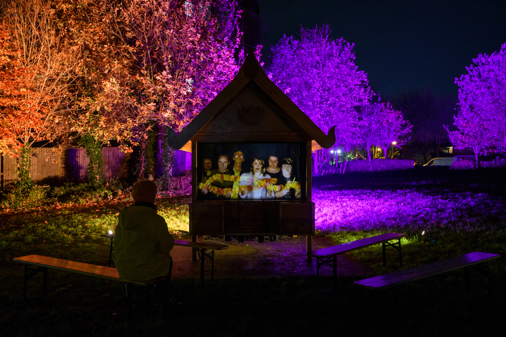 A person sits on a bence in a park lit up by purple and orange light, watching a video of a row of people holding flowers, inside a pitched-roof house structure.