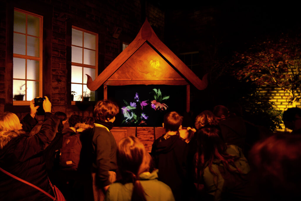 A group of people crowd around a video projection in a house-like wooden structure, set inside a darkened outdoor space beside a windowed brick wall. 