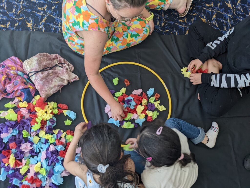 Three seated children and and adult photographed from above, arranging colourfuul paper petals inside a hoop. 