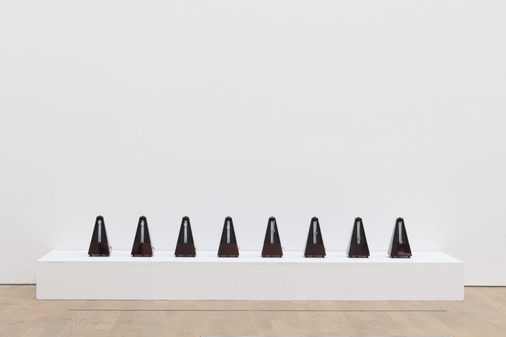 Eight black metranomes site on a low white plinth in a white walled gallery space.