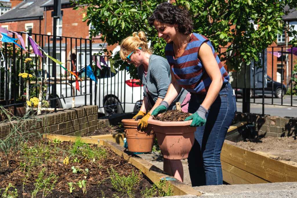 Two people hold pots full with compost. They are working on planting raised beds infront of them. The sun shines and they are smiling.