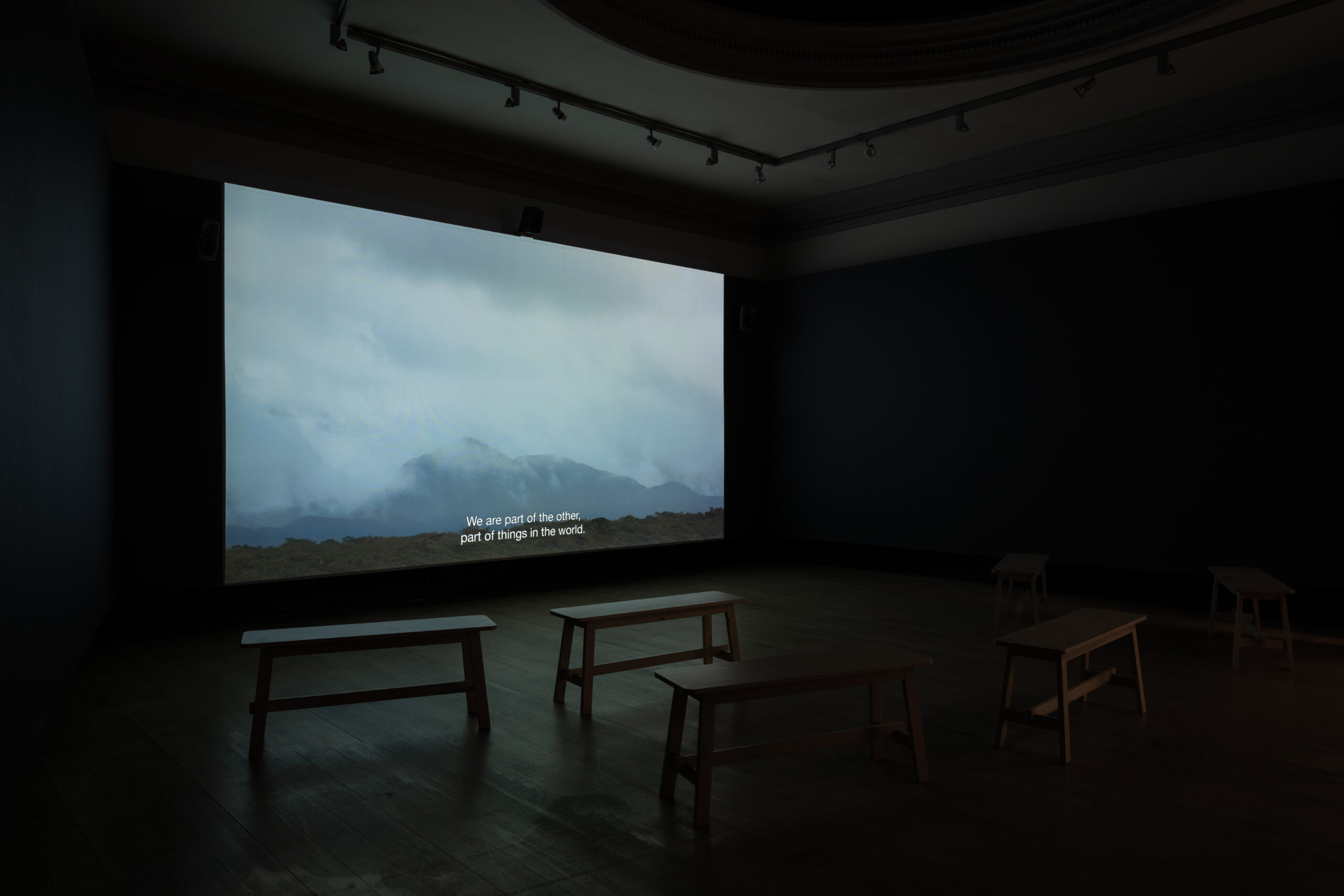 A dark gallery space with low benches arranged in front of a large screen showing a film still with a view of mountains in low cloud