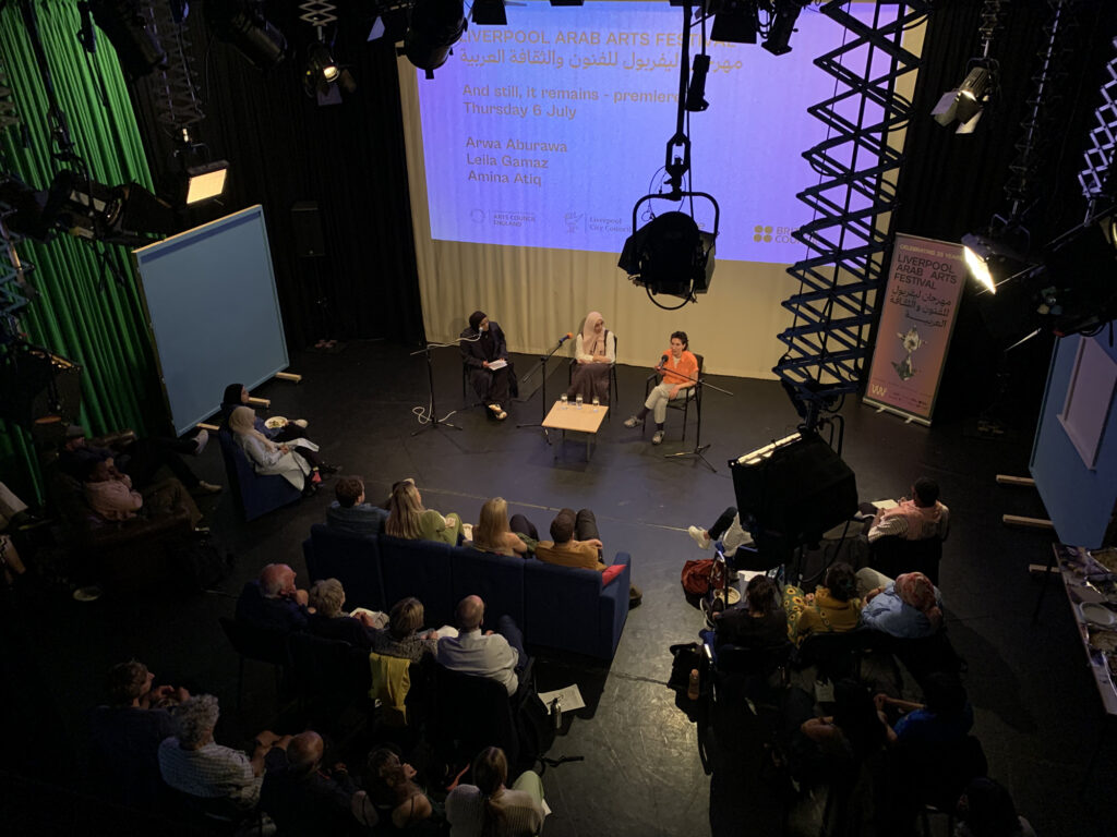 A shot from high up which looks down on an audience before a panel of three women with a large screen behind them
