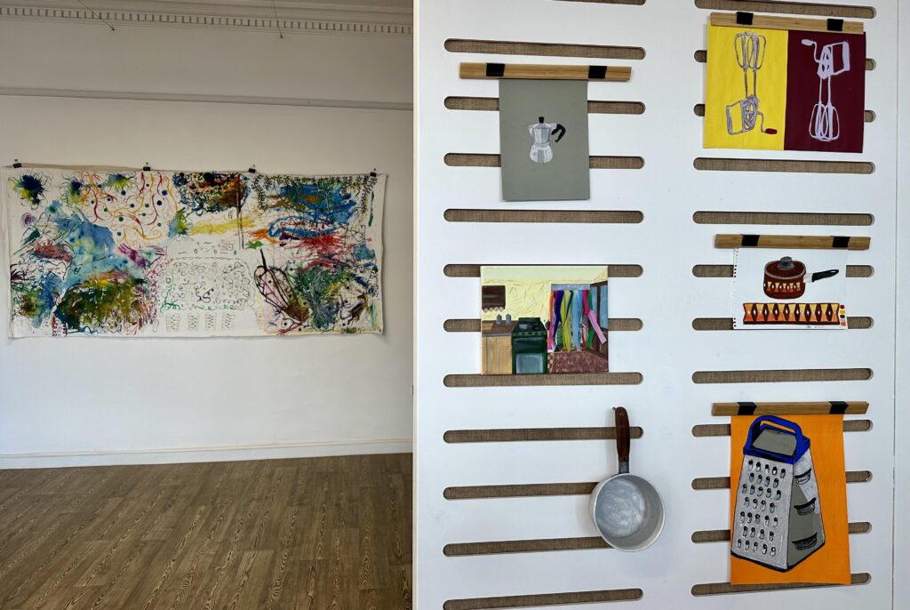 To the left is a wall set back with a large rectangular painting, abstract and multicoloured. To the right a wall in the foreground with 6 smaller pieces displayed