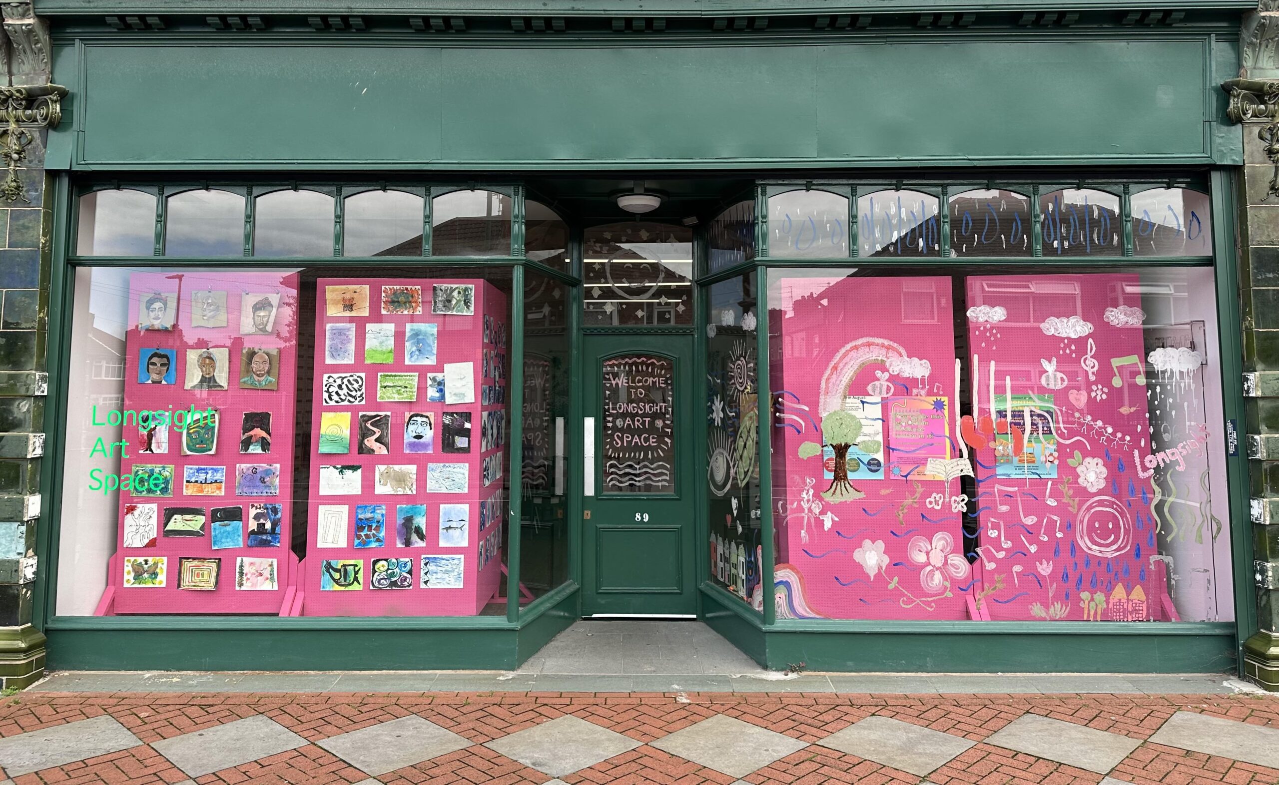The shop front has two big windows on either side and a recessed door in the middle. There are pink display boards with artworks in the windows