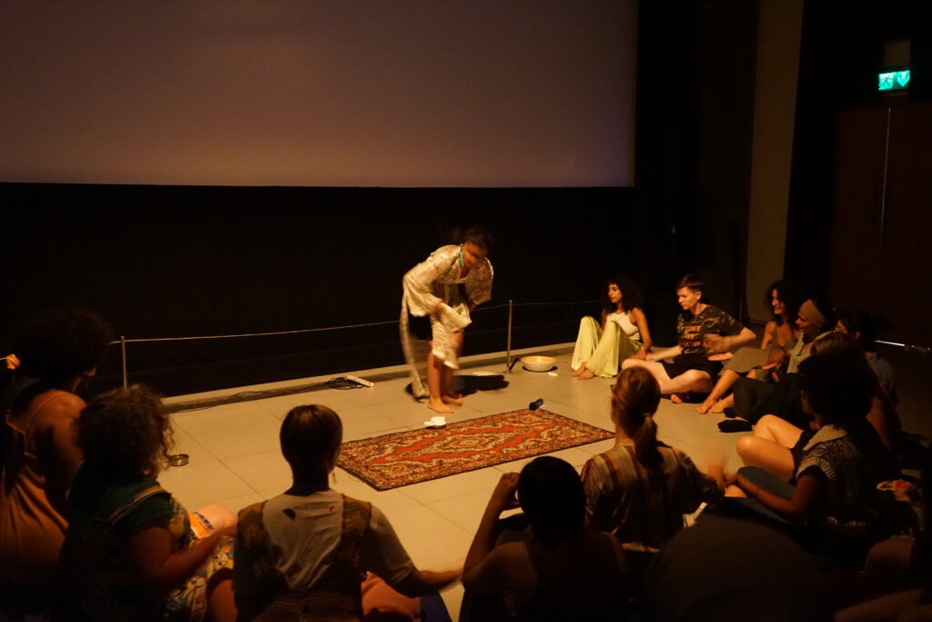 A group of people sit on the floor, cross legged, in a semi circle. Their arms are captured in motion, clapping together. In the centre of the semi circle a woman stands directing the group.
