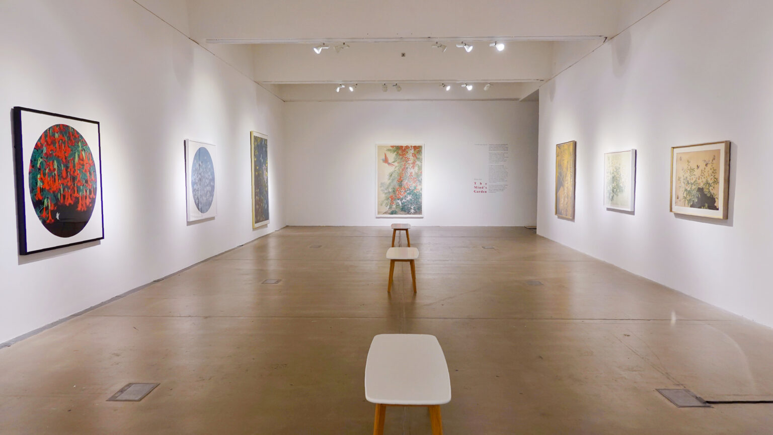 A white walled gallery space with a polished concrete floor. The view of the space encompasses three walls, there are three paintings on the left and right walls and one large painting on the wall facing. There are three benches in the centre of gallery space.
