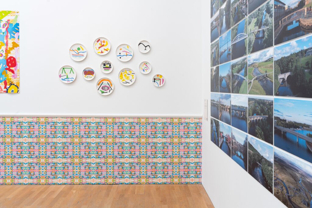 Gallery space with photographs of bridges on the right-hand wall, decorative plates and wallpaper on the back wall.
