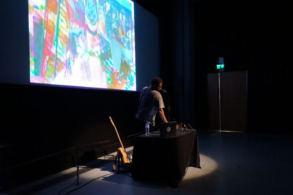 A man with brown skin and dark curly hair stands behind a table with electronic music equipment. He is performing. Behind him is a guitar and a cinema screen projecting an abstract, multi coloured collage image.