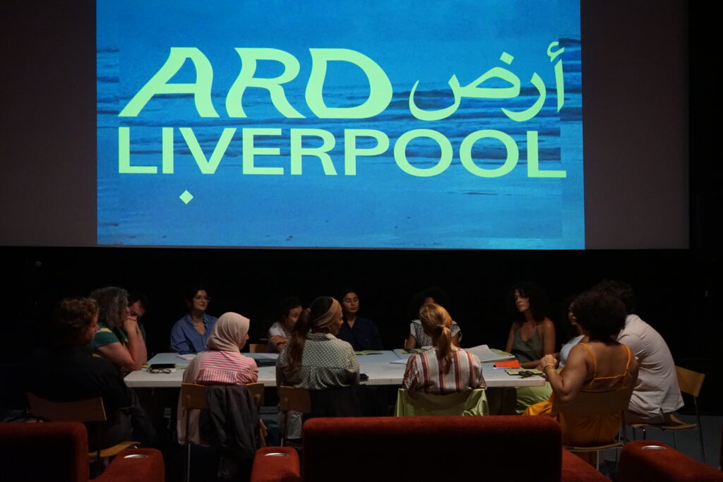 Fourteen people sit around a table. They all have texts (books, papers, phone screens) in front of them from which they are reading and discussing together. Behind them a cinema screen projects the words 'ARD Liverpool'.