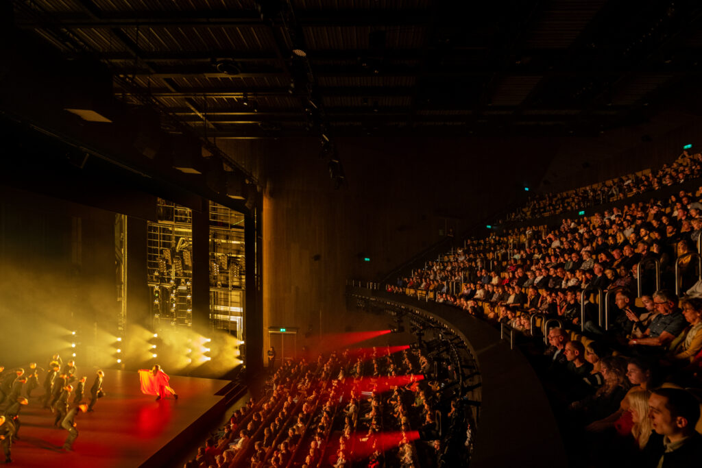 A side view inside the theatre which shows a full house with the circle and stalls visible, red and yellow spotlights pointing left towards the stage where dancers are visible