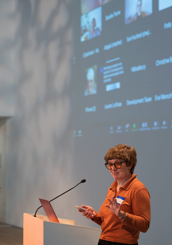 A person with short, thick ginger hair wearing an orange jersey top is stood at a podium in front of a large projection of a busy zoom meeting. She wears tinted glasses and is holding a remote control.