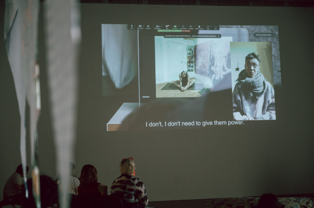 A view of a a group of people faving a large wall with a film made up of layered images projected onto it. Images include a woman in a yoga pose and a person facing the camera. The subtitle reads "I don't, I don't need to give them power.