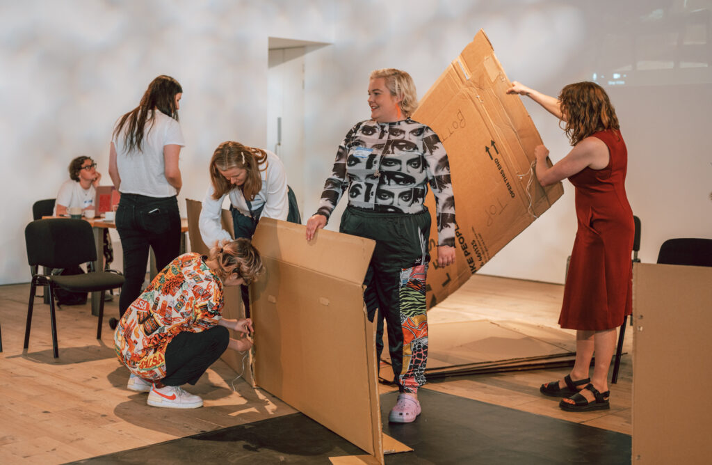 A group of six people are engaged in activity holding large sheets of cardboard and tying them together.