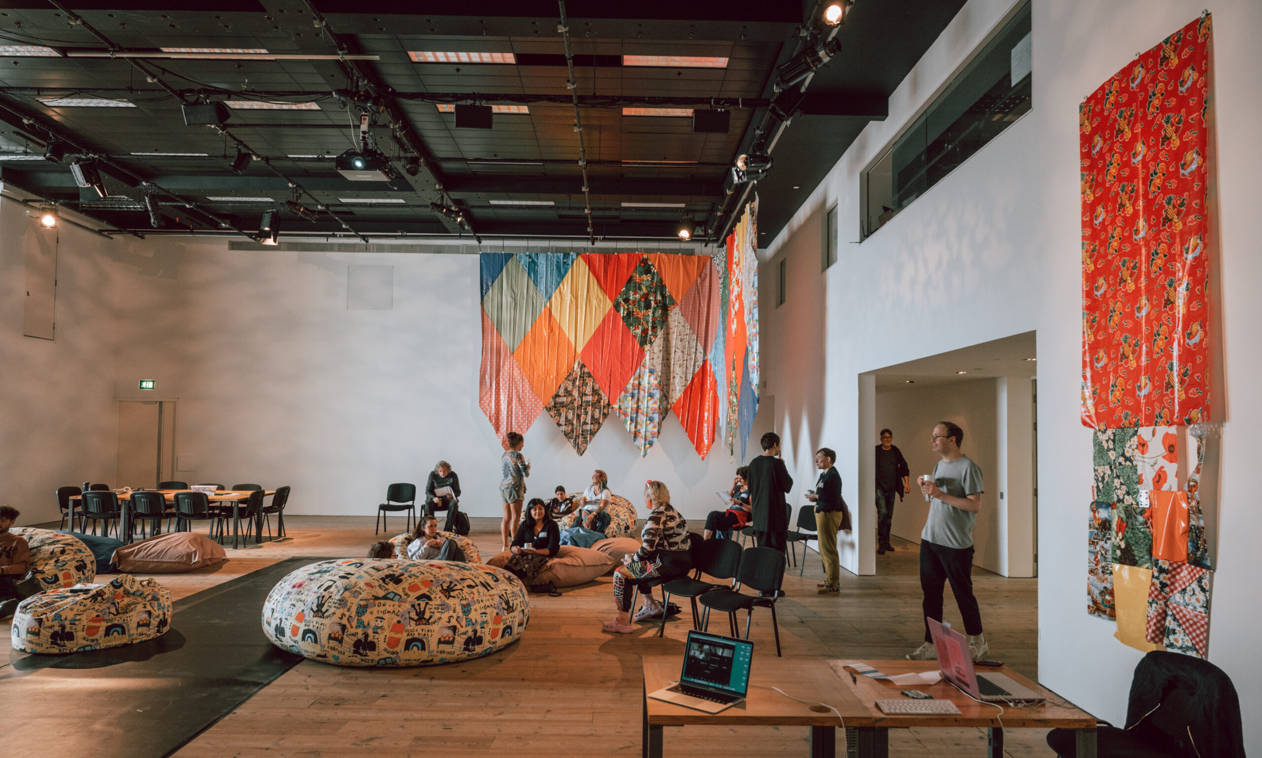 A large space with white walls and wooden floor, the ceiling is rigged with lights. There are a number of large beanbags on the floor, some have people sat in them. There is a large table at the back of the room with ten or so chairs around it. People mill around the space. Hanging on the back wall is a large patchwork hanging, large diamonds of coloured fabric stitched together. Another more simple square designed hanging is on the wall to our right.