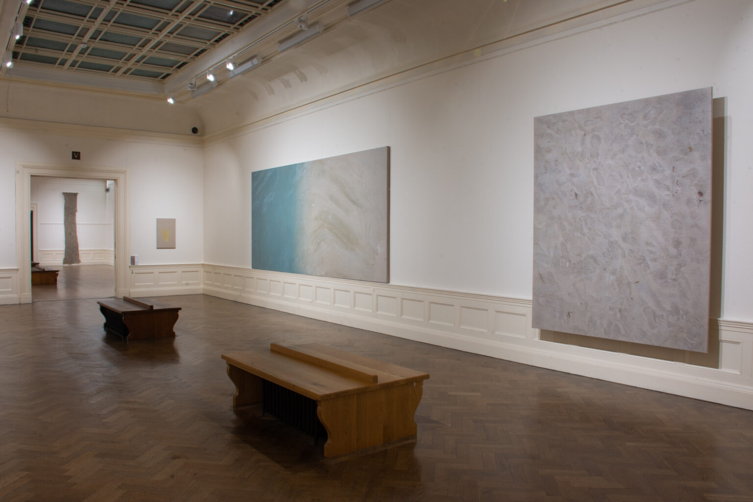 A white-walled gallery space with brown parquet flooring and two llow viewing benches. On the wall are two large canvases, on the left a gradient from blue to grey-beige, and on the right a mottled light brown.
