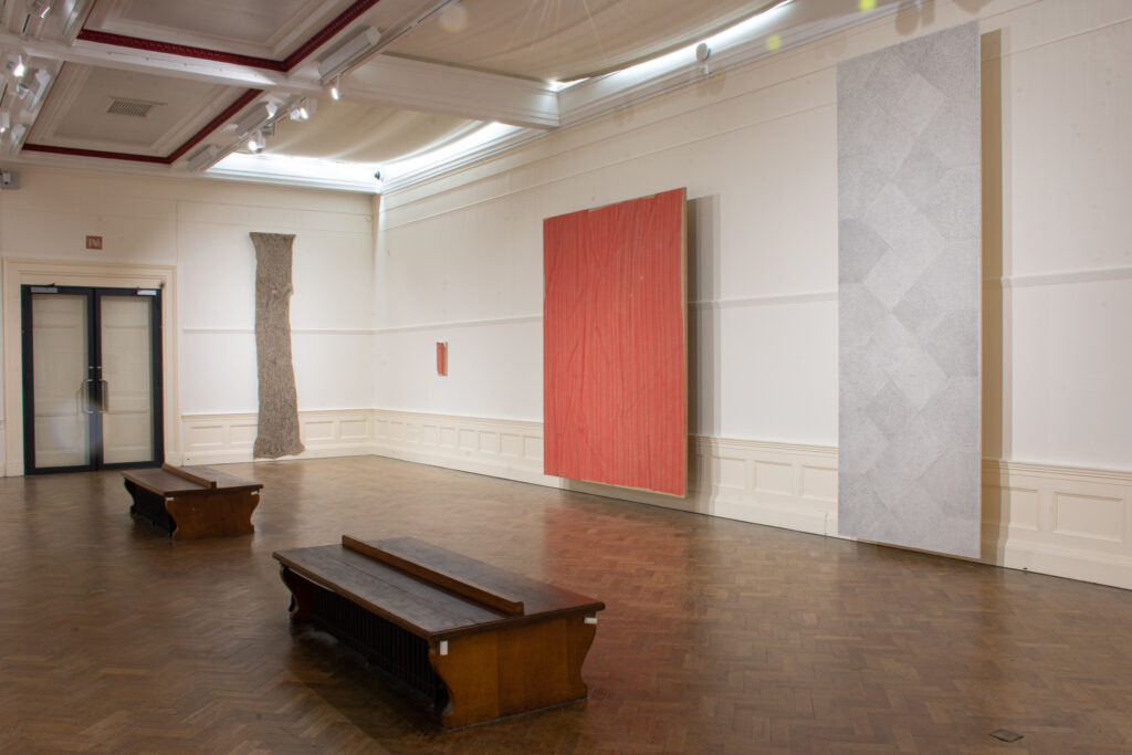 Three paintings are hung in the gallery space, on the left a long thin brown piece with irregular edges, in the middle a pink/orange work and on the right a tall piece, almost floor to ceiling, in pale grey.