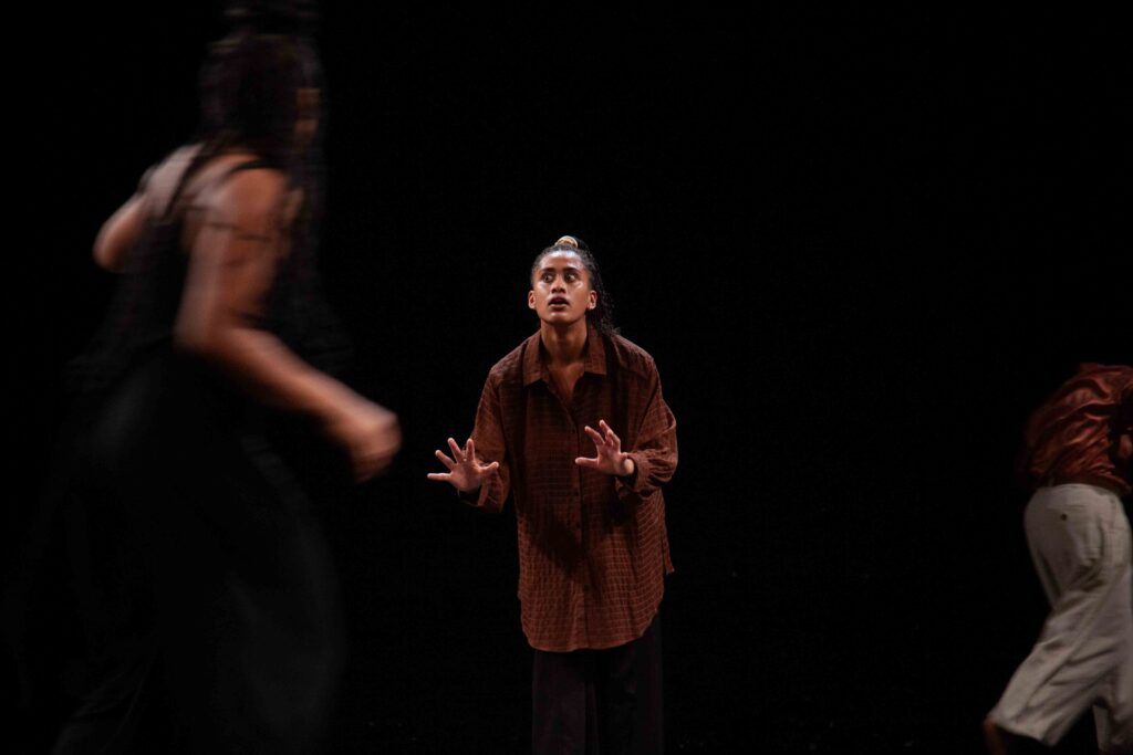 Three performers against a black background, the two people on either side blurred by their movements, the person in the middle static, eyes wide and hands outstretched as if about to touch something