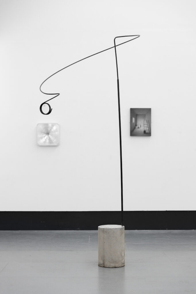 A sculpture, about five feet high, consisting of a steel rod twisted into a loop, set in a cylindrical block of cement sits on a light grey gallery floor. The wall behind is white and had two images on it, around A4 in dimensions.