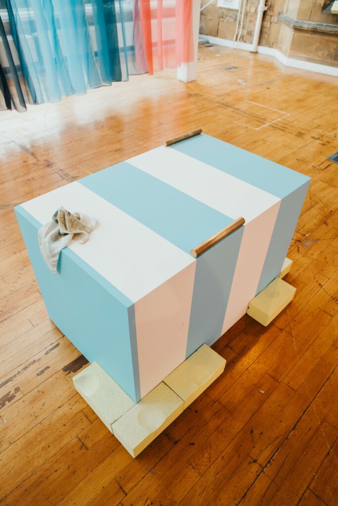 Blue and white striped box surrounded by yellow brick-like forms. On top of the box is a dirty hanky and two lengths of bronze rod.