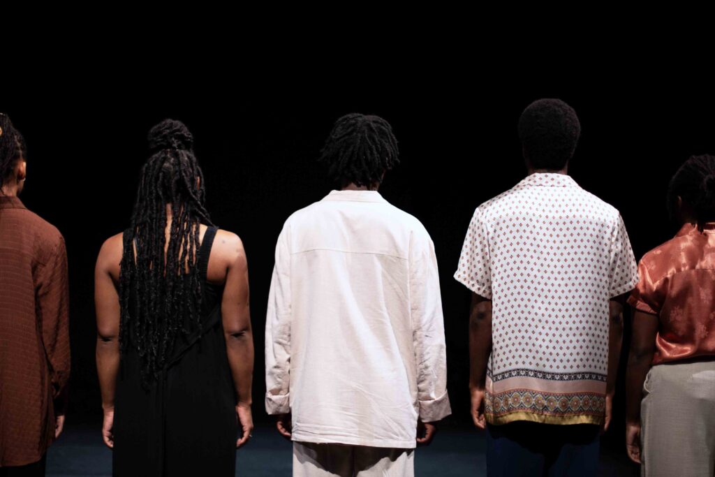 Five performers stand with their backs to the audience against a black background. Left to right they wear a brown, black, white, white with polkadots and a shiny orange shirt.