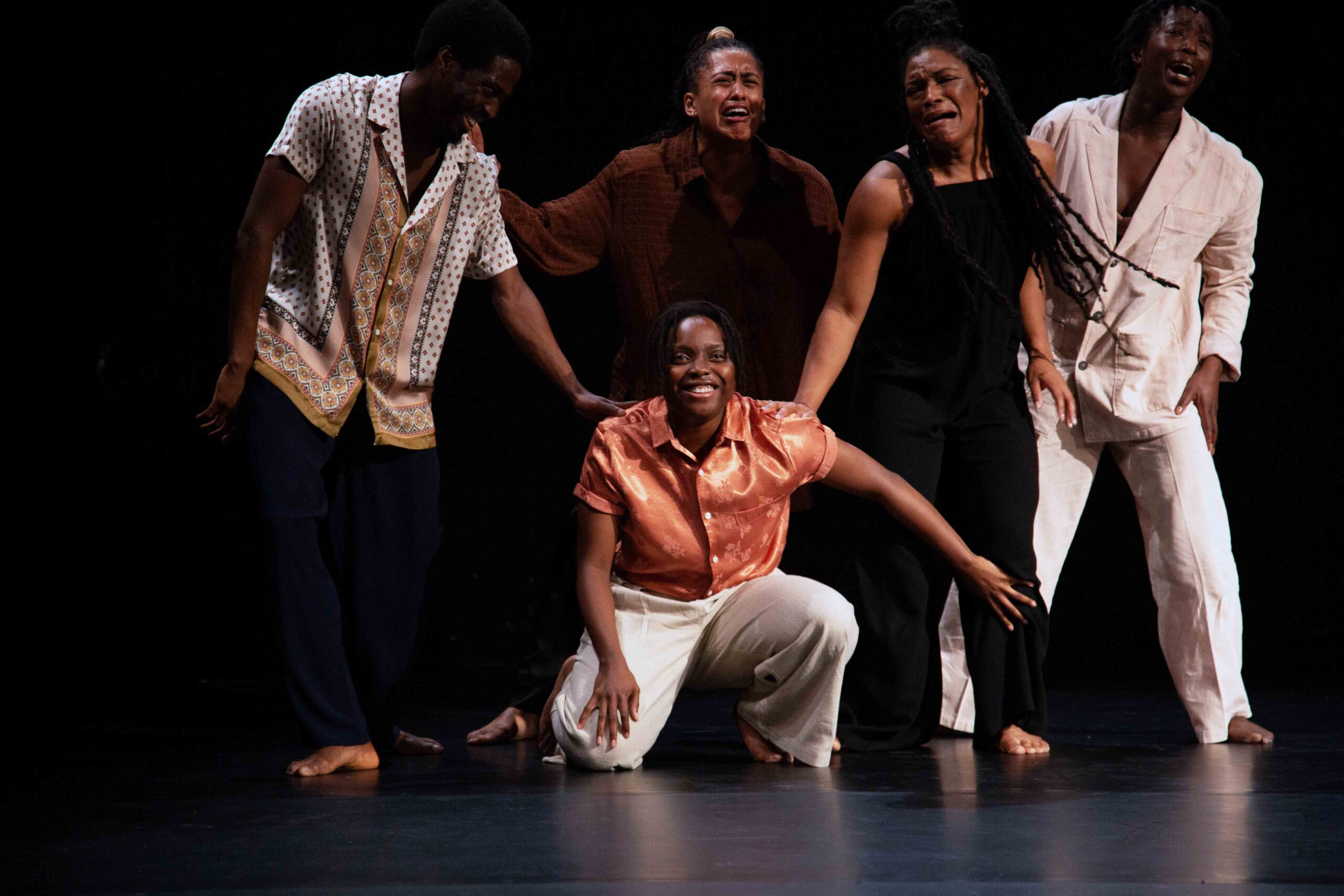 The five performers captured moving together, four upright with pained expressions on their faces, they seem to be crying, and a fifth performer in light orange shirt is kneeling on the floor in front of them.
