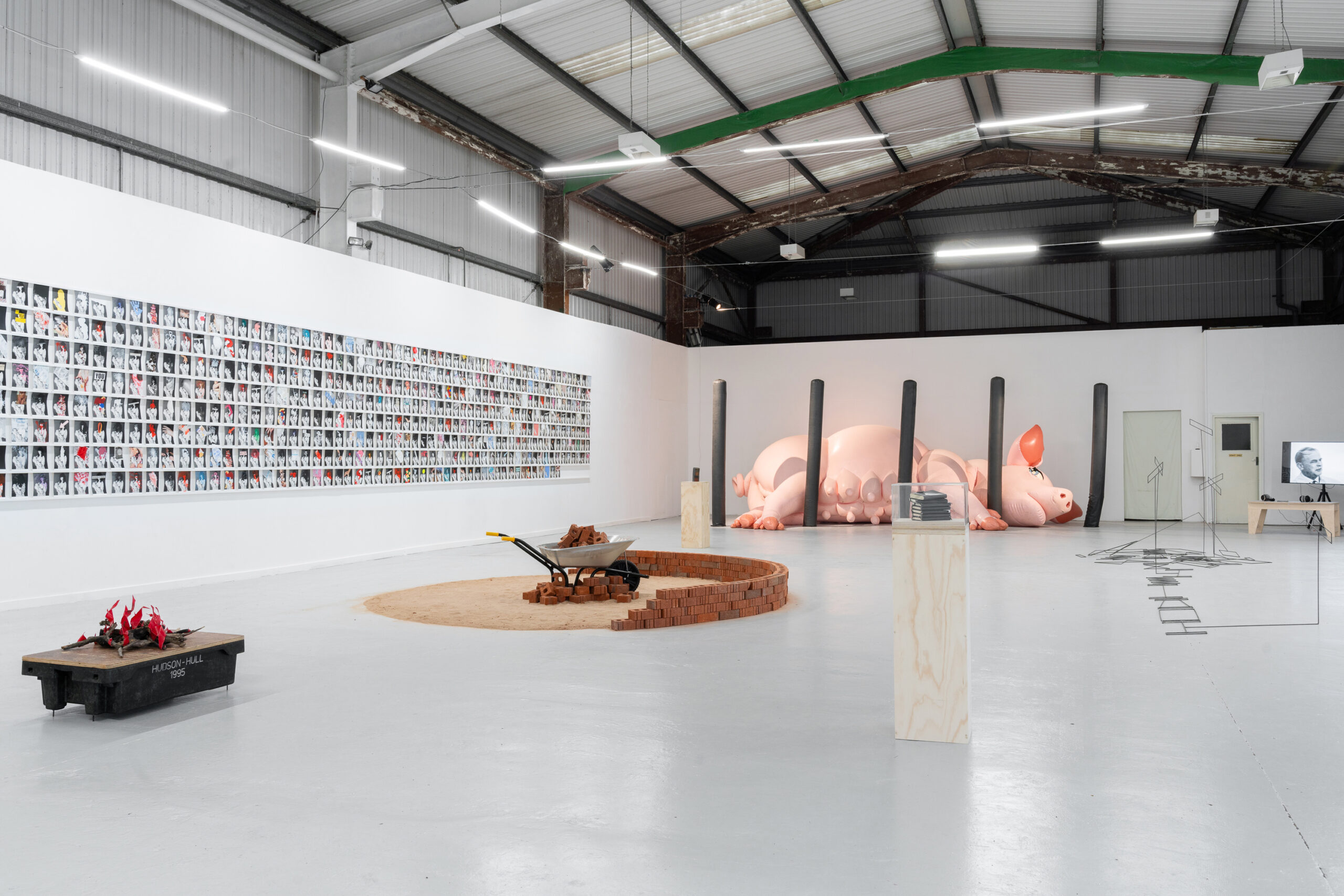 A large hangar like space with white walls and a light grey floor. In the foreground a box-like sculpture with plastic flames. Behind that a partially built circular wall and in the background a large inflatable pig laying on its side. On the wall to the right hundreds of small images displayed in a block.