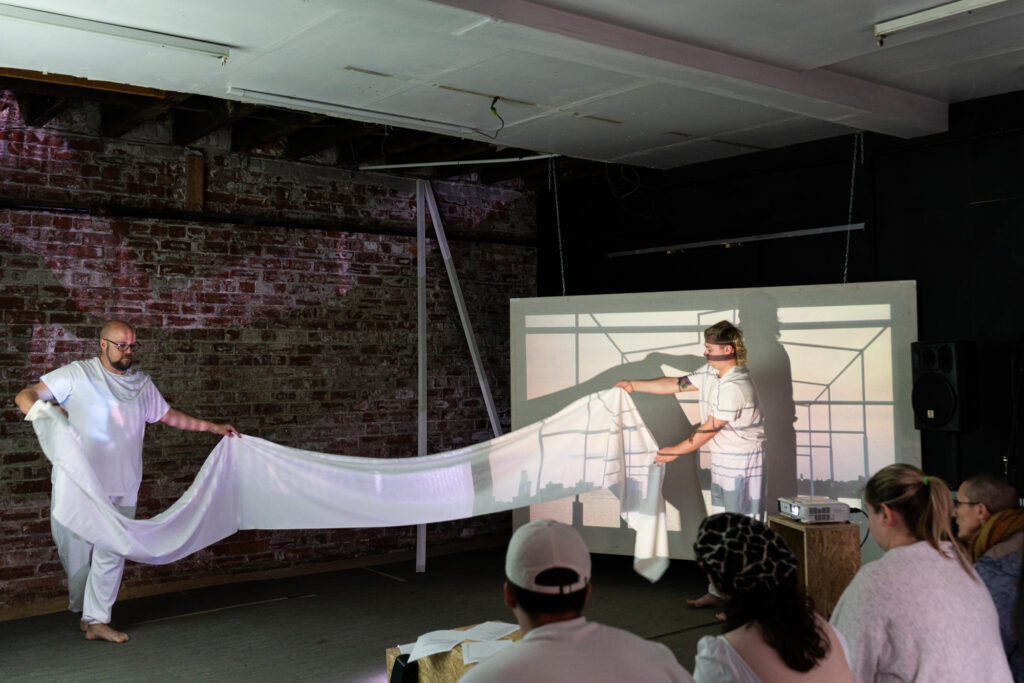 Two pale skinned people dressed in white t-shirts and pants are holding a length of white fabric, about three meters away from each other. Behind them is a brick wall and a white screen with an image of a building frame projected on. there are four people in the audience watching.