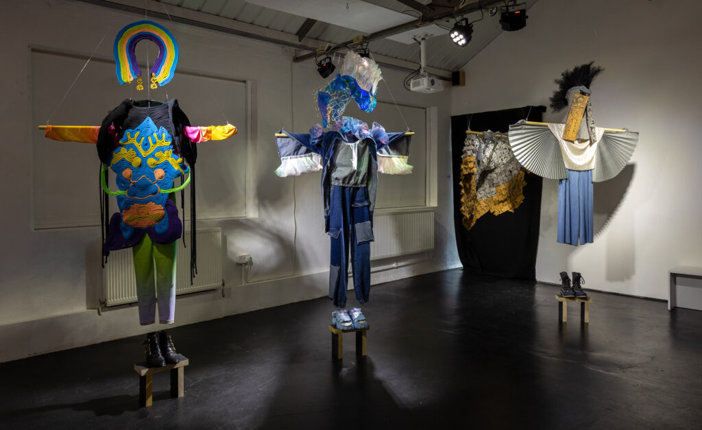Four colourful outfits hang in a dark-floored, white-walled gallery space.