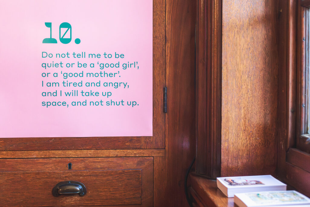 Text is printed in green on a pink background, the poster is mounted on a wood-panelled wall. The text says ''Do not tell me to be quiet, or a good girl, or a good mother, I am tired and angry and I wiull take up space, and not shut up"