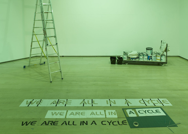A white walled gallery space lit up green, on the floor in the foreground stencils for lettering reading WE ARE ALL IN A CYCLE and to the left in the back a ladder, to the right a trolley with buckets of paint