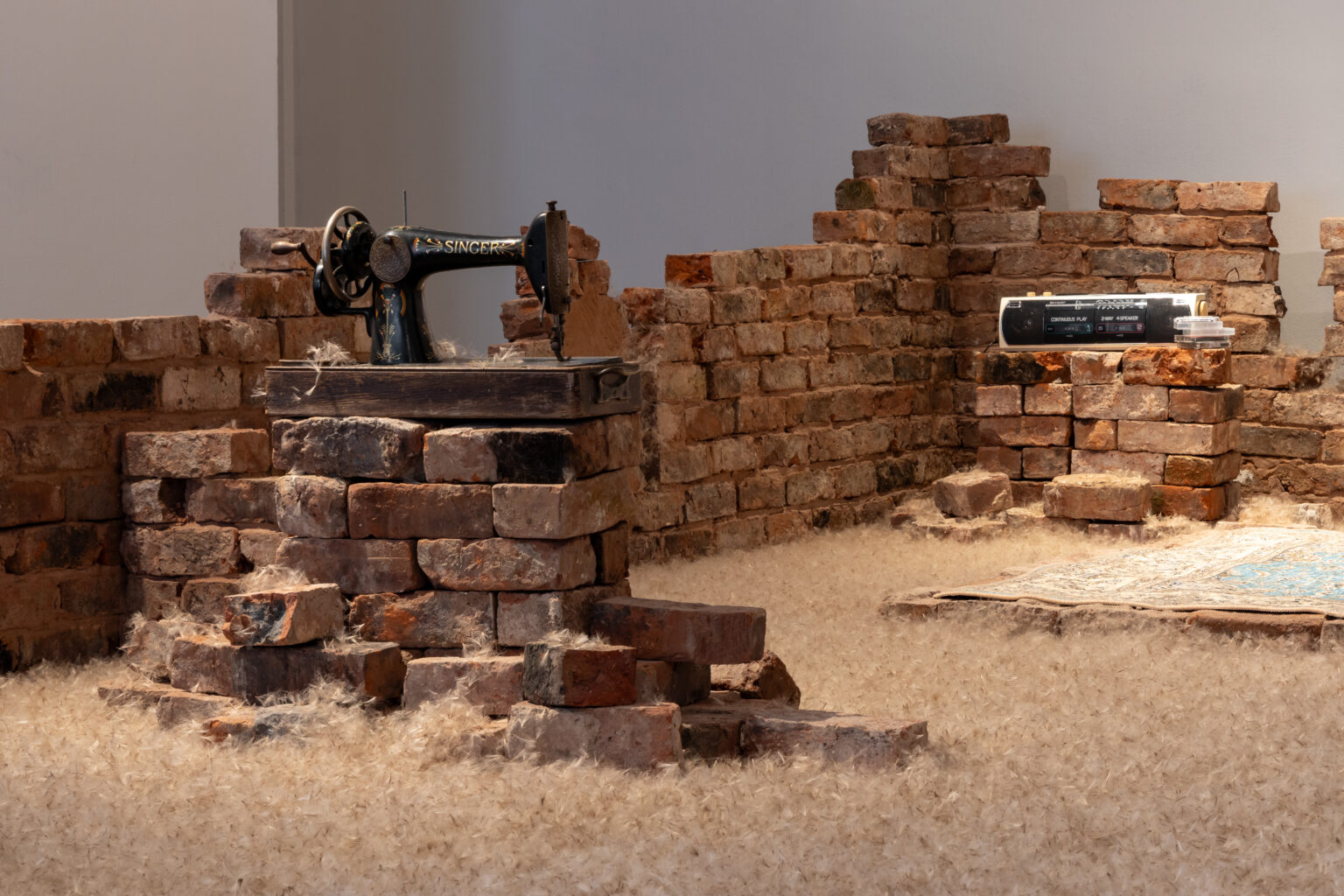 Low brick walls are build to different heights in a state of unfinishedness. There are two piles of bricks: one one an old black singer sewing machine, on the other an old tape player and stack of tapes. On the ground fluffy dandelion seeds like a carpet, and to the right an Iranian rug.