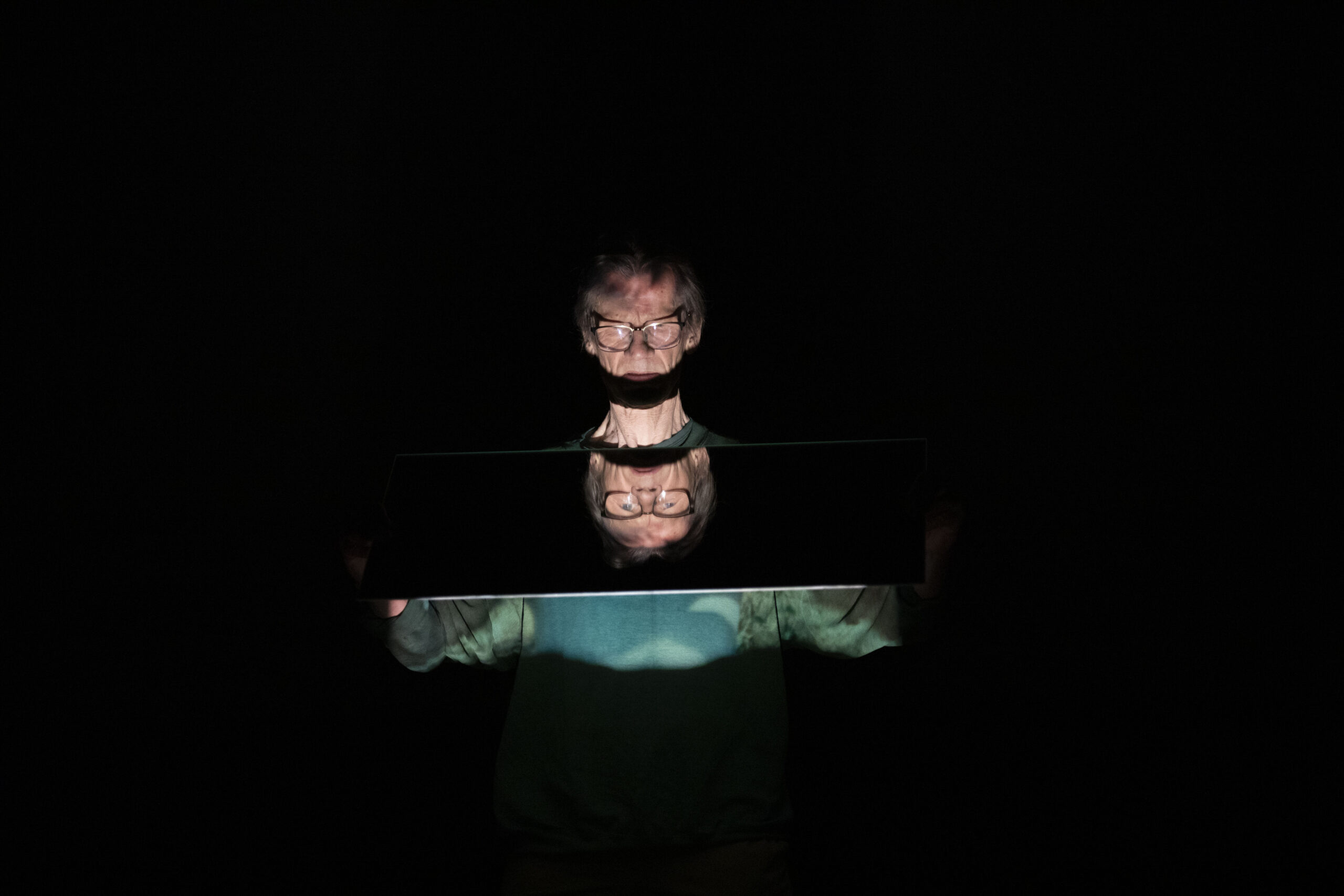 An older man with a pale complexion, grey hair and glasses, holds a mirror under his chin. The man's face reflected upside-down, doubling his head