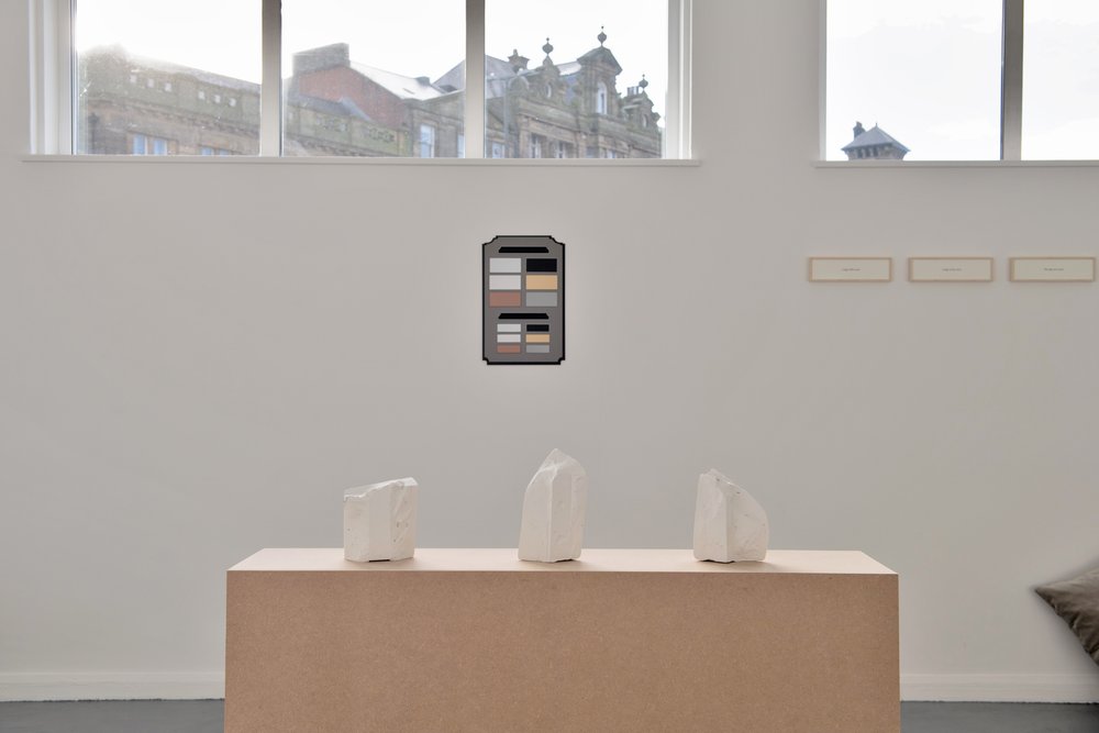 In the foreground is a wide beige plinth with three small plaster objects arranged in a row. On the wall behind is a grey plaque, about A4 in size, with a number of small colourd rectangles displayed on it. Above the plaque the windows show the view of rooftops outside.